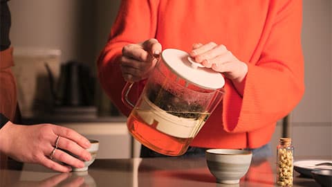Someone wearing an orange jumper pouring from the Teapot REEDesigned. There are two grey cups on the counter and the plunger in the teapot is being pressed down and the tea leaves at the top of the water.